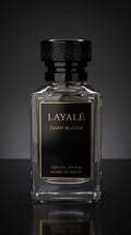 A clear rectangular glass perfume bottle with a black cap and a black front label featuring gold lettering that reads 'LAYALÉ DARK BLOOM'. The bottle is centered against a dark gray gradient background with a reflective surface below.