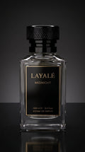 A clear glass perfume bottle with a black textured cap and a black front label featuring gold lettering that reads 'LAYALÉ MIDNIGHT'. The bottle is centered against a dark gray gradient background with a reflective black surface below.