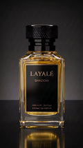 A rectangular glass perfume bottle filled with amber liquid, featuring a black cap and a black front label with gold lettering that reads 'LAYALÉ SHADOW'. The bottle is centered against a dark gray gradient background with a reflective surface below.